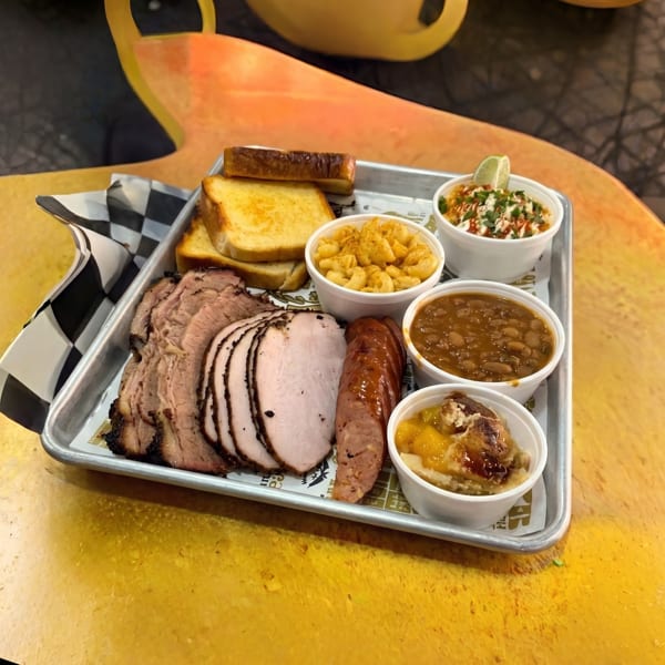 A metal tray holds sliced brisket, turkey, sausage, toast, mac and cheese, baked beans, and peach cobbler.