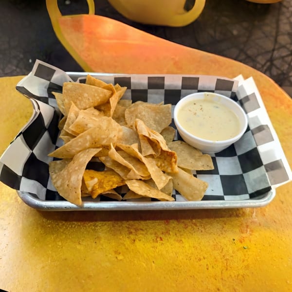 A metal tray with a checkered paper liner holds tortilla chips and a cup of queso dip.