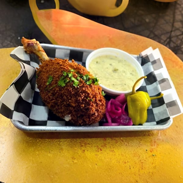 Crispy fried chicken drumstick with green sauce, pickled red onions, and a yellow pepper on a tray.