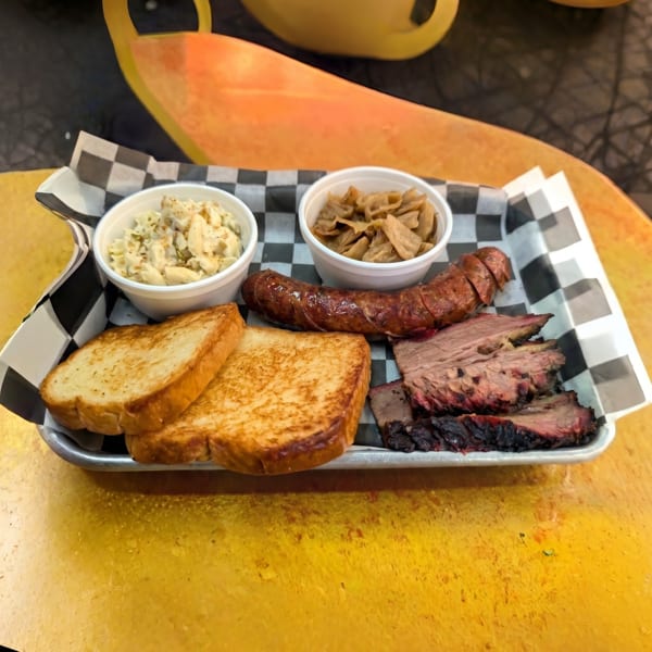 A BBQ platter with sliced brisket, sausage, toast, macaroni salad, and stewed cabbage.
