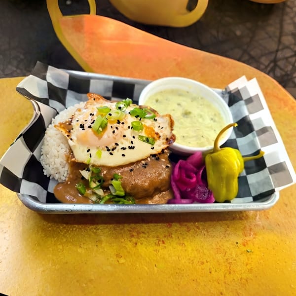 Loco Moco with a fried egg, gravy, rice, pickled onions, and a pepper on a metal tray.